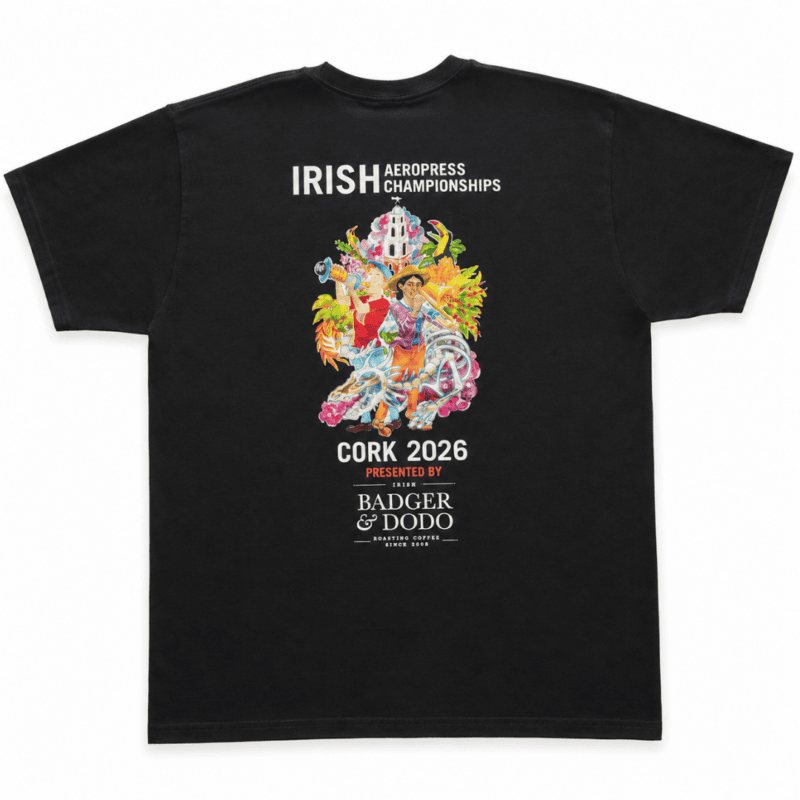 Back of a black T-shirt featuring colorful artwork of people, a church, tropical flowers, a dragon, and a coffee cup, with white text reading “Irish Aeropress Championships,” “Cork 2026,” and “Presented by Badger & Dodo Roasting Coffee.