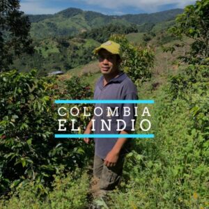 A man wearing a yellow cap and dark polo shirt stands among coffee plants on a lush hillside in Colombia, with green mountains and cultivated farmland in the background. Text overlay reads “COLOMBIA EL INDIO.”