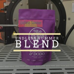 A purple coffee bag labeled “Endless Summer Blend” by Kafex & Dodo sits on a metal outdoor table in front of a large coffee roasting machine. White overlay text reads “ENDLESS SUMMER BLEND,” with yellow horizontal lines above and below the title.