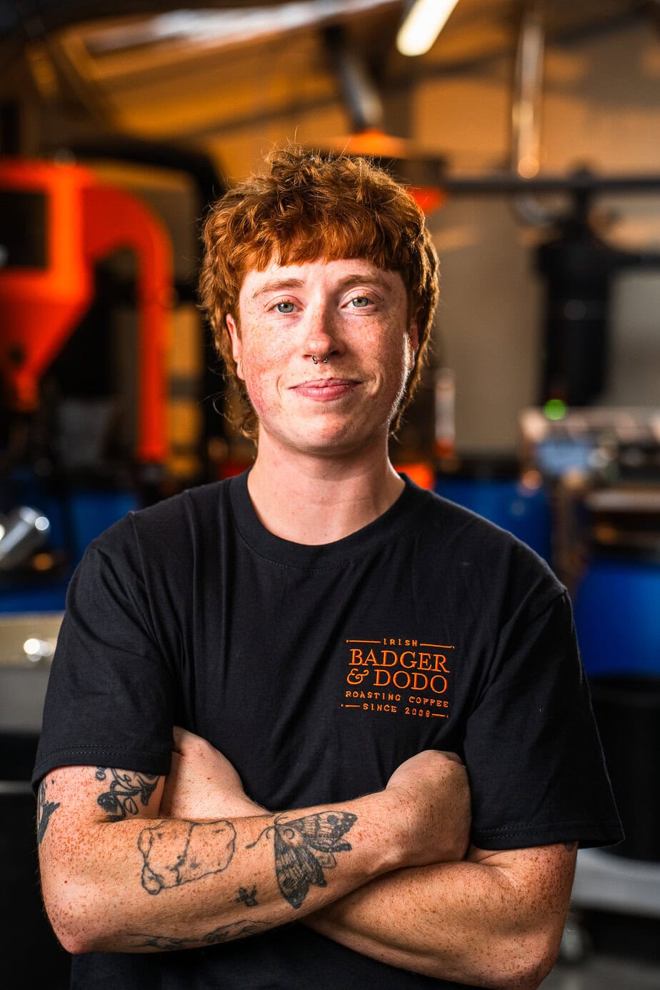 Person with curly red hair standing in a grassy field, wearing a black “Badger & Dodo” t-shirt and black shorts with a white drawstring. They are smiling with arms crossed, showing tattoos on their forearms. The sky is cloudy in the background.